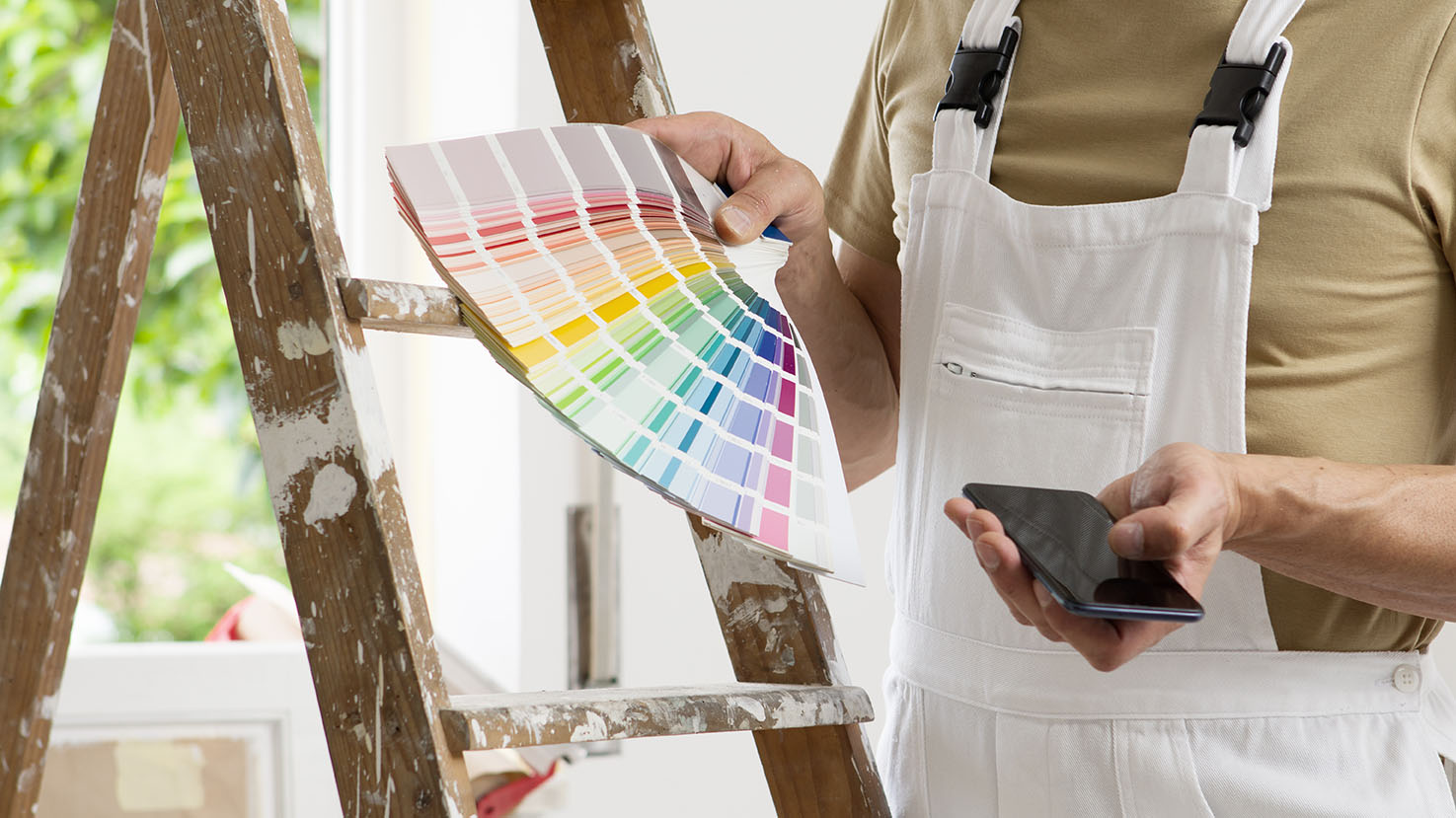 hands of house painter man decorator choose the color using the sample swatch, work of the house to renovate, use mobile phone, a wooden ladder and a green window as a background, close up